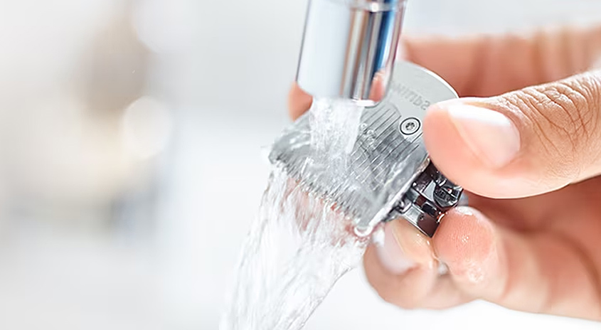 Eine Hand hält einen silbernen Trimmer unter einen Wasserhahn, aus dem klares Wasser fließt, vor einem weißen Hintergrund.