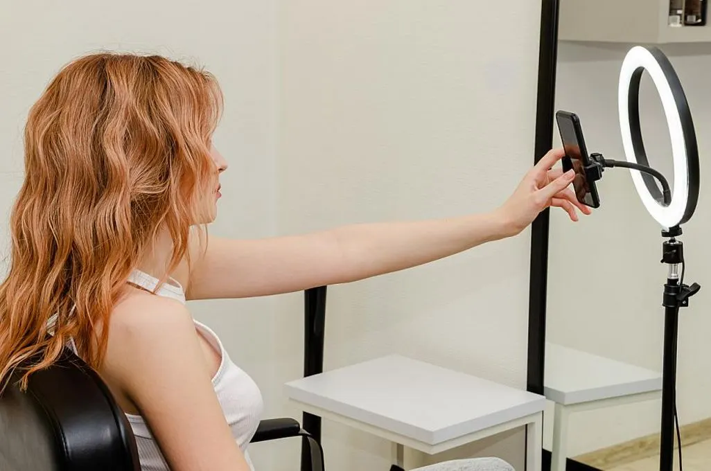 Frau sitzt auf einem Stuhl und richtet ihr Smartphone in einer Halterung mit Ringlicht aus.