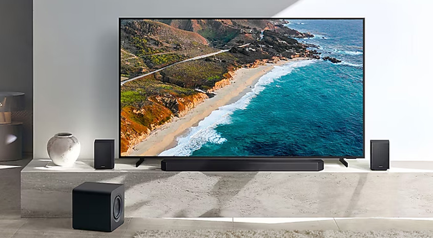 Ein Fernseher zeigt eine Küstenlandschaft. Darunter befinden sich eine Soundbar, zwei kleine Lautsprecher und ein Subwoofer auf einem grauen Podest.
