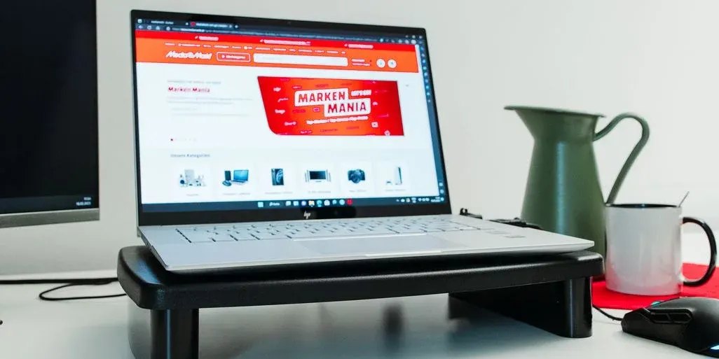 Silberner Laptop auf schwarzem Ständer mit MediaMarkt-Website. Links dunkler Monitor, rechts grüne Kanne, weiße Tasse, schwarze Maus auf roter Matte.
