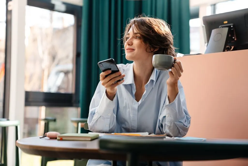 Frau sitzt im Café und hält Smartphone und Kaffeetasse.
