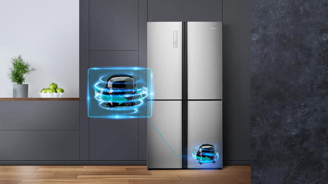 Ein viertüriger, silberner Kühlschrank steht auf einem Holzboden vor einer grauen Wand. Eine Pflanze steht auf einer Fensterbank.