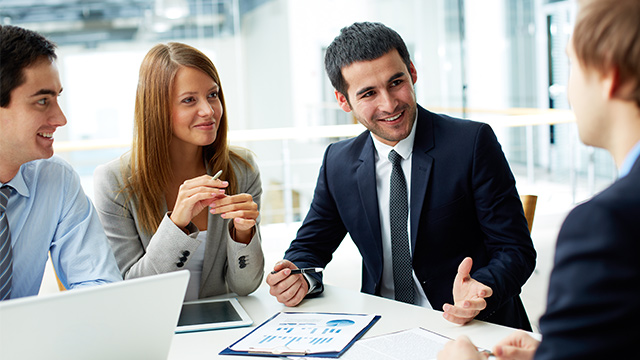 Eine Gruppe von vier Personen sitzt an einem Tisch mit einem Laptop, einem Tablet und Diagrammen in einem hellen Büro.
