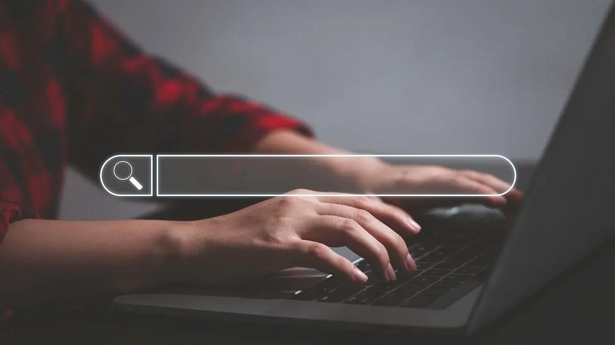 Hände tippen auf Laptop-Tastatur. Ein weißes Suchfeld mit Lupensymbol ist eingeblendet. Im Hintergrund: Person in rot-schwarz kariertem Hemd, graue Wand.