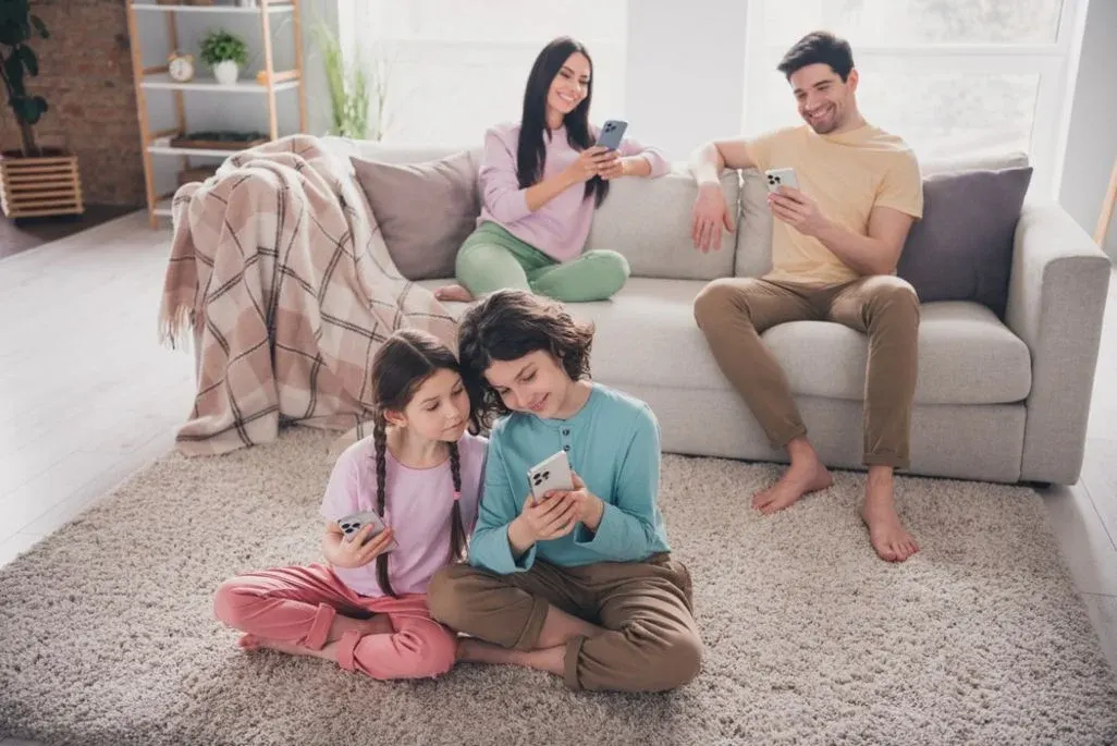 Zwei Kinder sitzen im Wohnzimmer am Teppich und schauen gemeinsam auf ein Handy, im Hintergrund sitzen 2 Erwachsene mit Handy auf einer beigen Couch