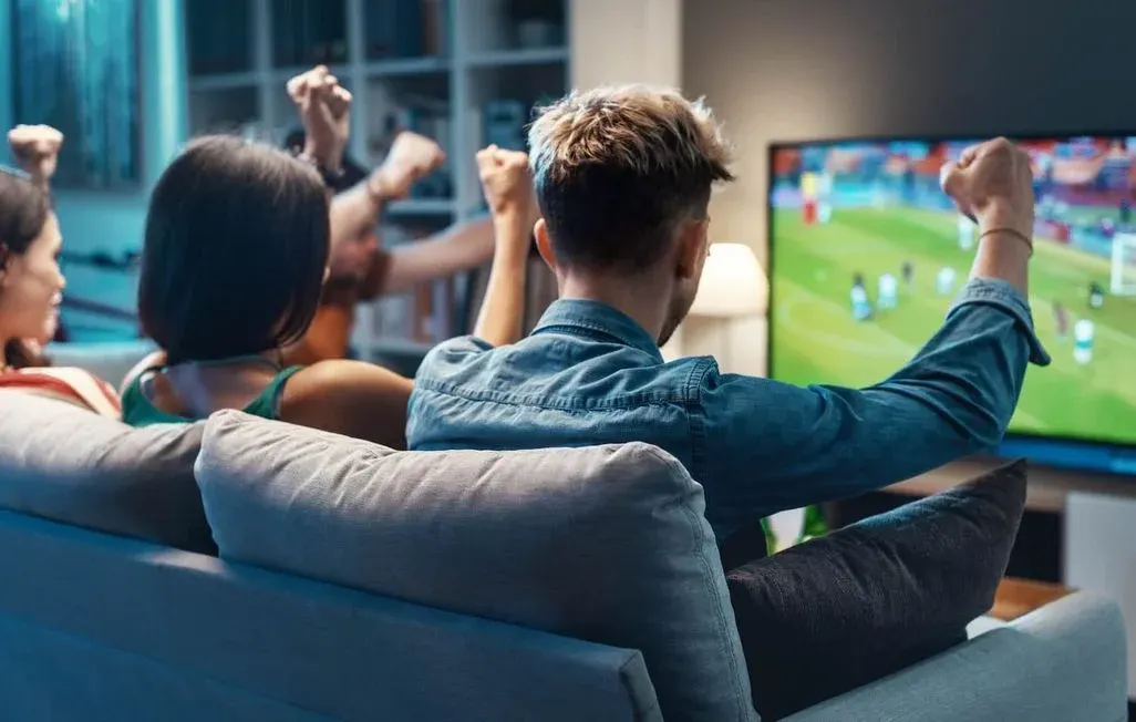 Gruppe von Freunden sitzt auf Sofa und jubelt vor Fernseher. Fußballspiel läuft auf dem Bildschirm. Gemütliches Wohnzimmer mit modernem TV-Setup.