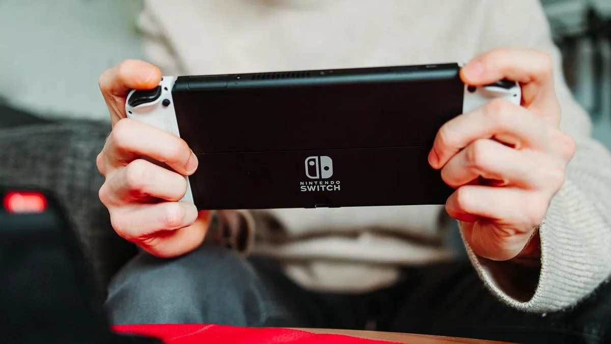 Hände halten schwarze Nintendo Switch Konsole mit weißen Controllern; Logo sichtbar. Person sitzt über rotem Stoff.