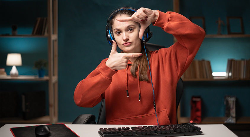 Eine Person mit Kopfhörern formt ein Rechteck mit den Händen vor einem Schreibtisch mit Computerzubehör. Bücherregale im Hintergrund.