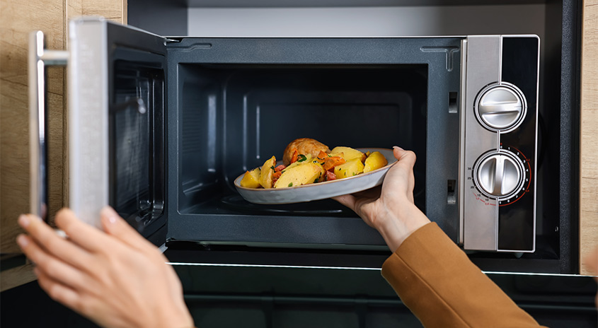  Person schiebt einen Teller mit heißen Kartoffeln und Gemüse in eine geöffnete Mikrowelle mit silberner Front und zwei großen Drehknöpfen.