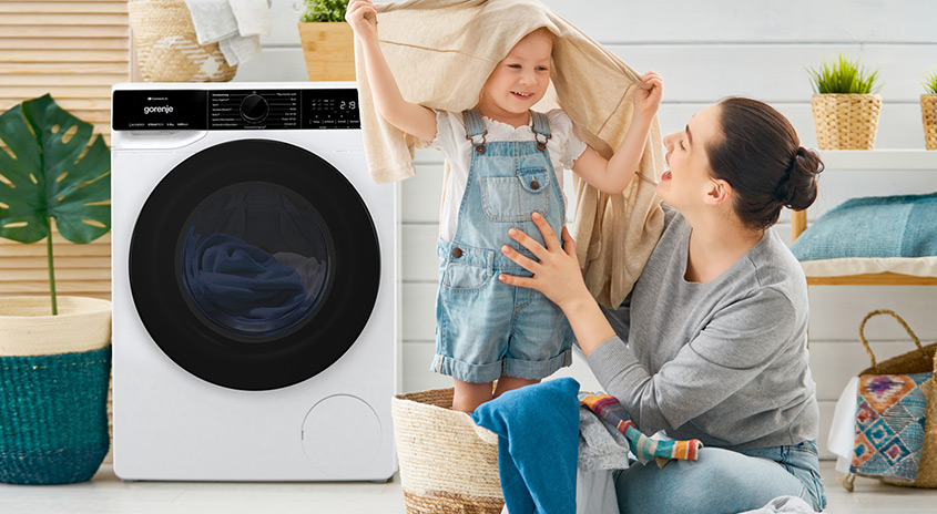 Eine Frau und ein Kind spielen mit Wäsche vor einer weissen Waschmaschine. Pflanzen und Körbe schmücken den Raum.