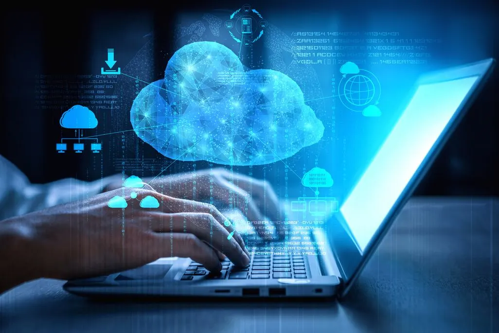 Hände tippen auf einem Laptop. Darüber schwebt eine große, leuchtend blaue Wolke mit digitalen Verbindungssymbolen und Datenströmen vor blauem Hintergrund.
