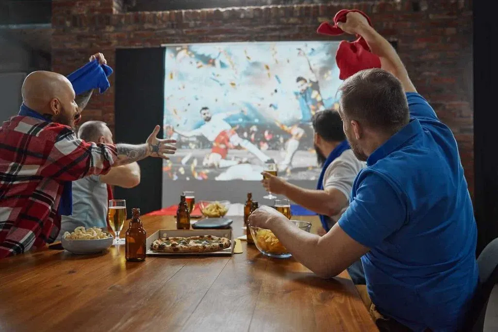 Vier Männer sitzen an einem Holztisch mit Pizza und Bier und jubeln begeistert mit erhobenen Armen, während sie ein Fußballspiel auf einer großen Leinwand schauen.