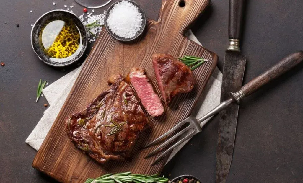 Ein geschnittenes Steak auf einem Holzbrett mit Rosmarin, Salz, Olivenöl, einem Messer und einer Gabel auf dunklem Untergrund.