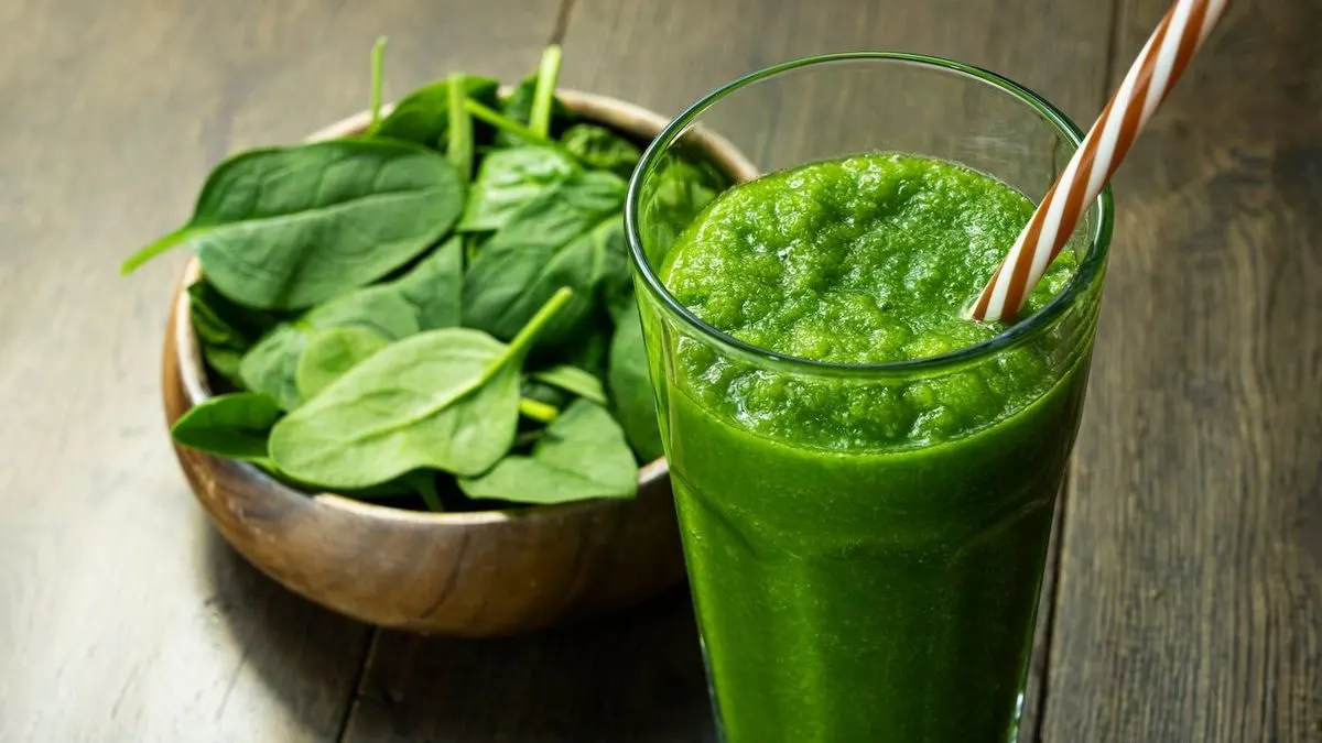 Ein Glas grüner Smoothie mit gestreiftem Strohhalm steht neben einer Holzschale voller frischem Spinat auf dunklem Holz.