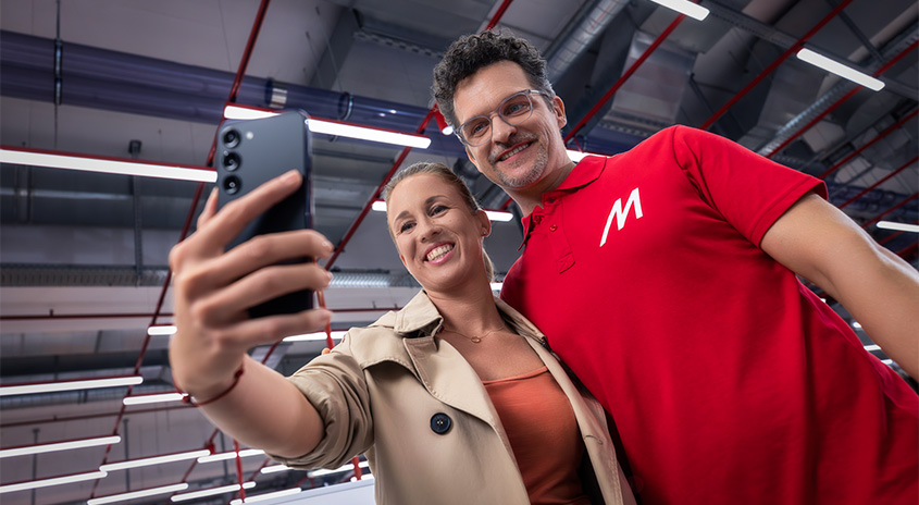 Eine Frau hält ein schwarzes Smartphone, um ein Foto zu machen. Ein Mann in einem roten Hemd steht daneben.