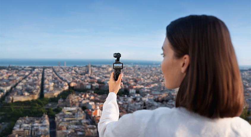 Frau mit weißem Hemd filmt Stadtpanorama mit Kamera-Gimbal. Weitblick auf Küstenstadt mit Meer, Häuser und blauem Himmel.