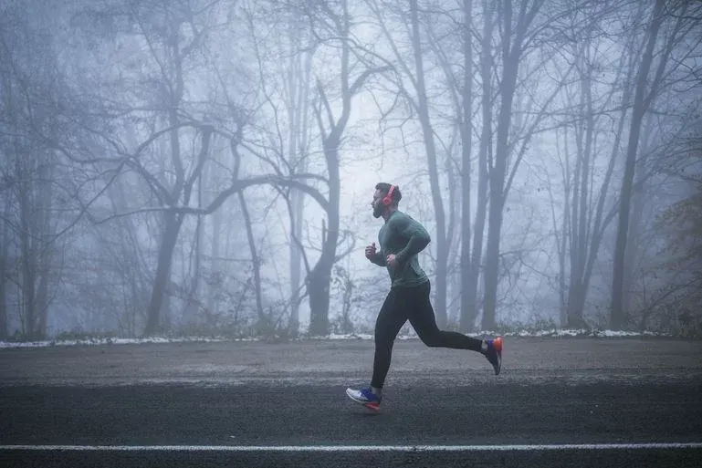 Mann mit orangefarbenen Kopfhörern, grünem Shirt und schwarzen Hosen läuft auf dunklem Weg. Kahle Bäume im dichten Nebel bilden den Hintergrund.