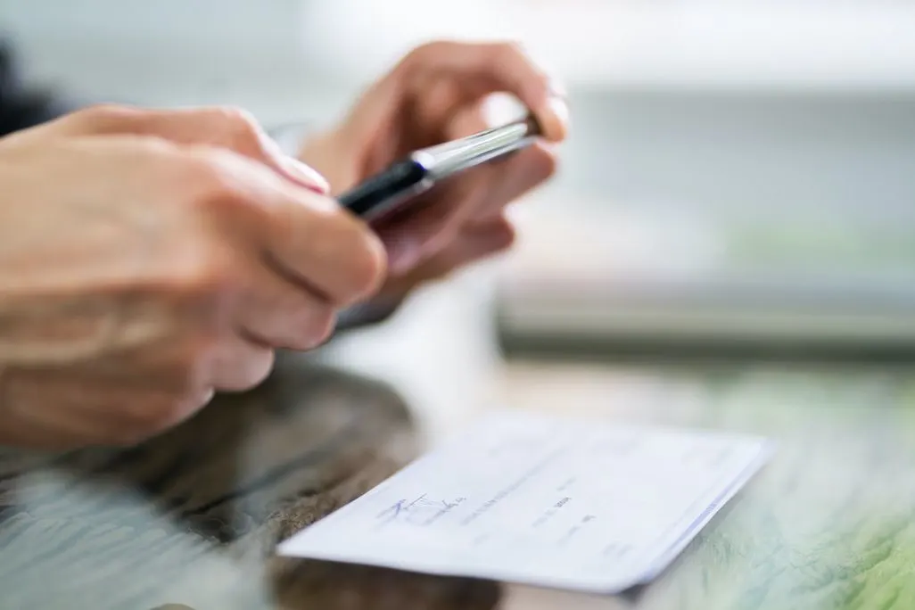Hände halten ein schwarzes Smartphone über einen weißen Scheck mit blauer Schrift, der auf einer Oberfläche liegt.