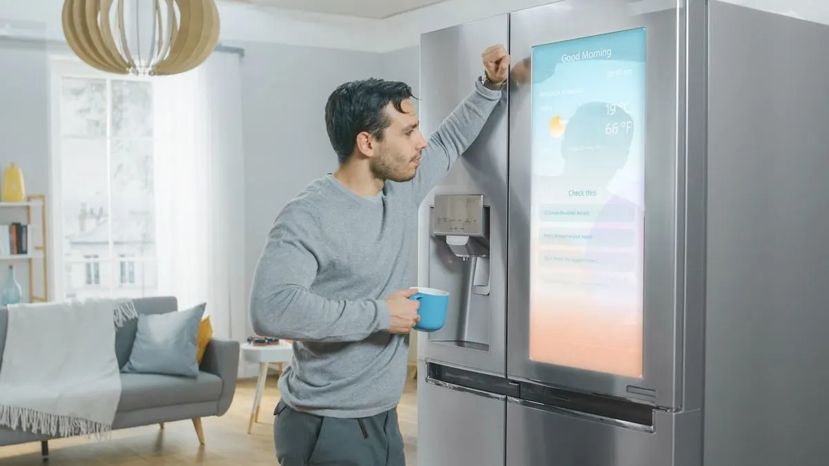 Mann in grauem Hemd mit blauem Becher lehnt an einem silbernen Smart-Kühlschrank. Der Bildschirm zeigt "Guten Morgen" und Wetterdaten. Ein Sofa und ein Fenster sind im Hintergrund.