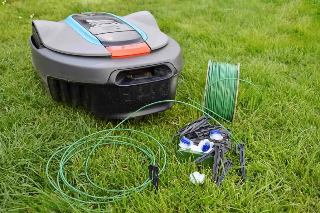 Rasenmähroboter auf einer Wiese mit ausgerolltem Begrenzungskabel, Kabelrolle, Heringen und Verbindungssteckern zur Installation.