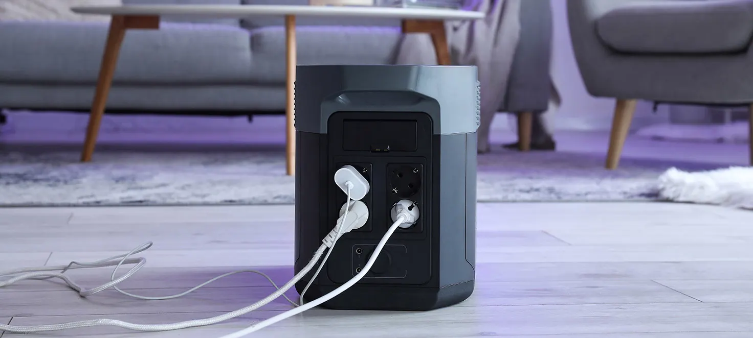 Ein graues Kraftwerk mit zwei weißen Netzkabeln steht auf einem Holzboden. Im Hintergrund befinden sich Möbel auf einem Teppich.