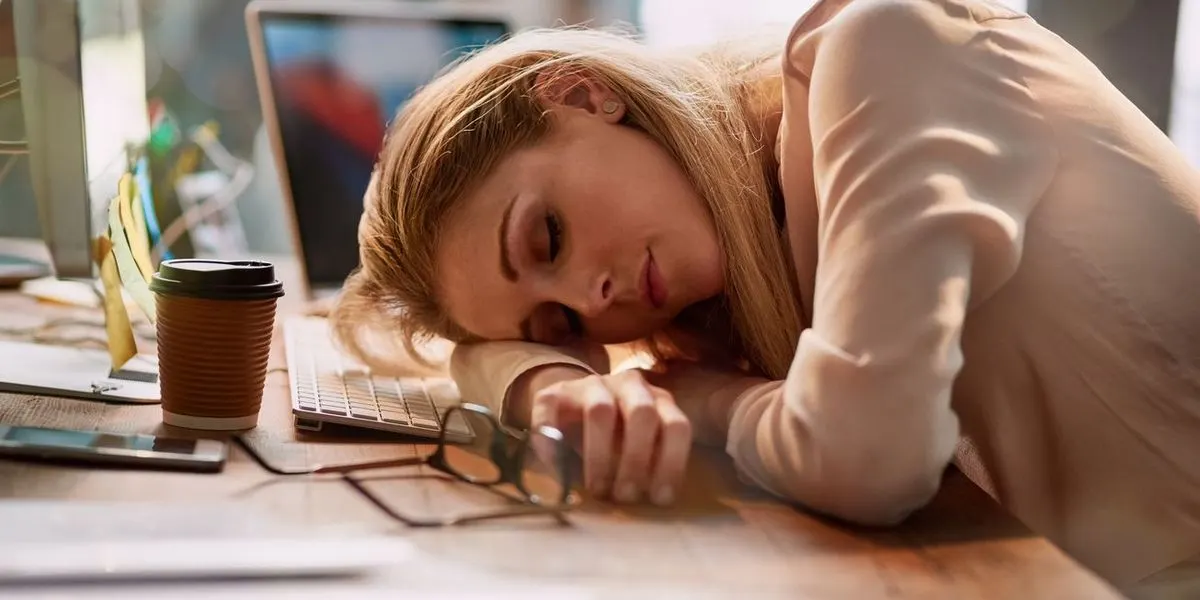 Frau liegt mit dem Kopf auf dem Arm an einem Schreibtisch mit Laptop, Tastatur, Brille und Kaffeebecher.