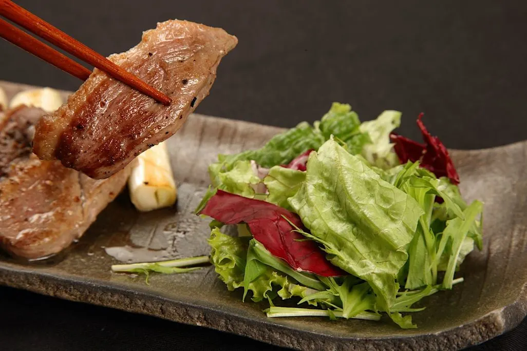 Gegrilltes Stück Fleisch wird mit Essstäbchen neben Salat gehalten.