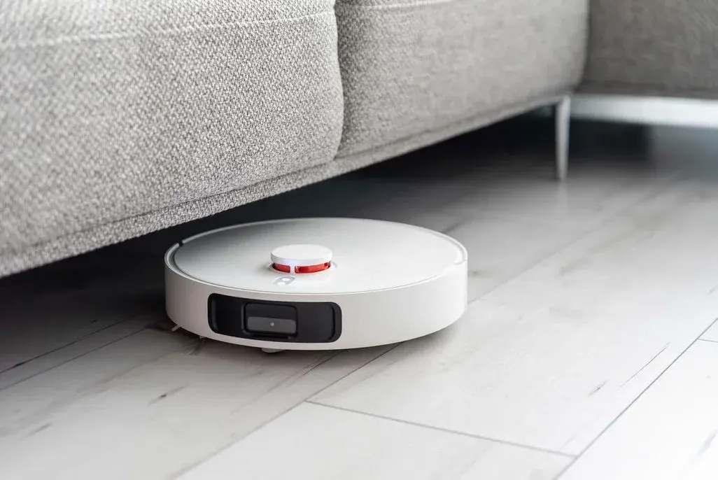Weißer Saugroboter mit rotem Sensor unter grauem Sofa auf hellem Holzboden.
