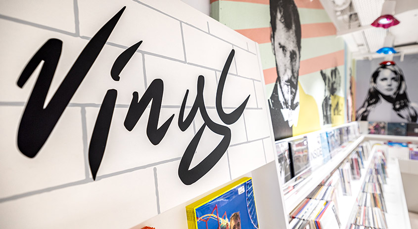 Schwarze Schrift "Vinyl" auf weißer Ziegelwand. Regale mit Schallplatten und Porträtgemälde im Hintergrund. Helle Deckenleuchten.