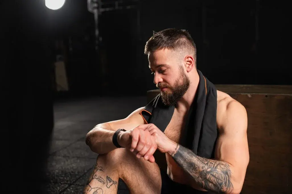 Ein bärtiger Mann mit schwarzem Handtuch und Fitness-Tracker sitzt auf dunklem Boden, lehnt an Holz und schaut auf sein Handgelenk. Tattoos sichtbar.