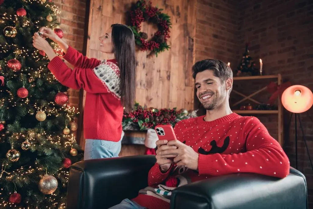 Frau dekoriert beleuchteten Weihnachtsbaum mit roten/goldenen Kugeln. Mann sitzt lächelnd im Sessel, nutzt rotes Handy. Festliche Dekoration.