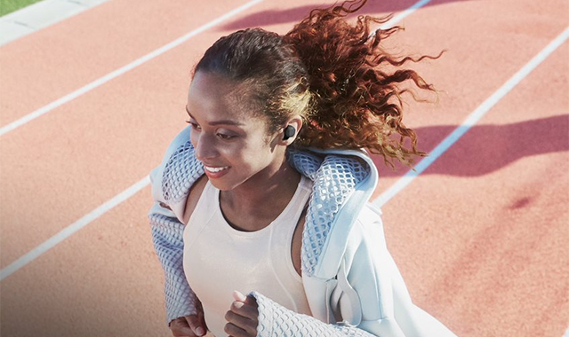Eine Frau mit einem weißen Tanktop und einer hellblauen Jacke läuft auf einer roten Laufbahn. Sie trägt Kopfhörer.