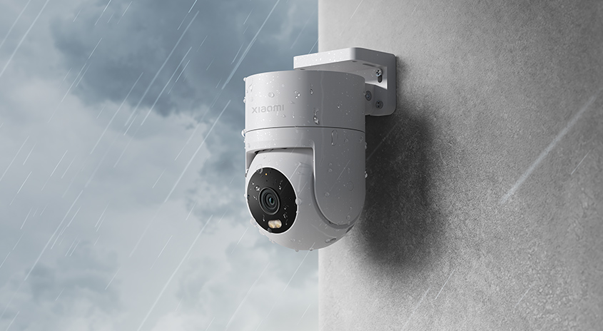 Eine weiße Überwachungskamera ist an einer grauen Wand im Regen befestigt. Der Himmel ist bewölkt.