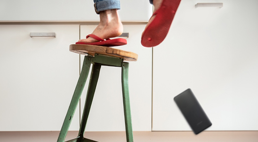 Auf dem Bild ist ein grüner Hocker mit einem Holzbrett als Sitzfläche zu sehen. Eine Person trägt rote Flip-Flops und ein Handy fällt herunter.