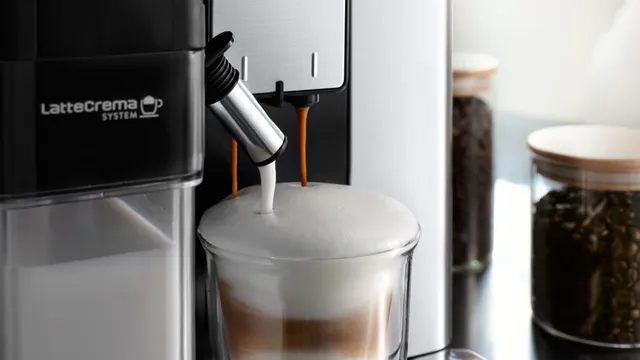 Nahaufnahme einer Kaffeemaschine, die Milchkaffee in ein Glas gießt. Im Hintergrund stehen Kaffeebohnen in Gläsern.