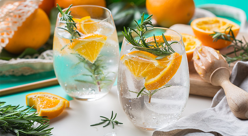 Zwei Gläser Sprudelwasser mit Eis, frischen Orangenscheiben und Rosmarinzweigen auf Tisch, umgeben von ganzen und halbierten Orangen sowie Holz-Zitruspresse.