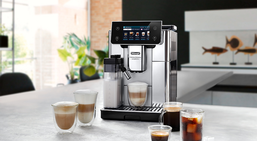 Eine silberne Kaffeemaschine steht auf einem Tisch mit mehreren Gläsern Kaffee in einem hellen Raum.