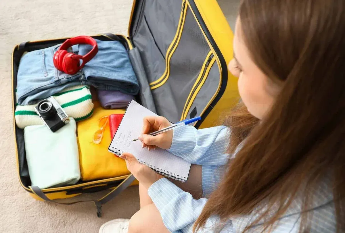 Frau sitzt vor einem geöffneten und fertig gepackten Koffer und notiert eine Packliste für den Urlaub.