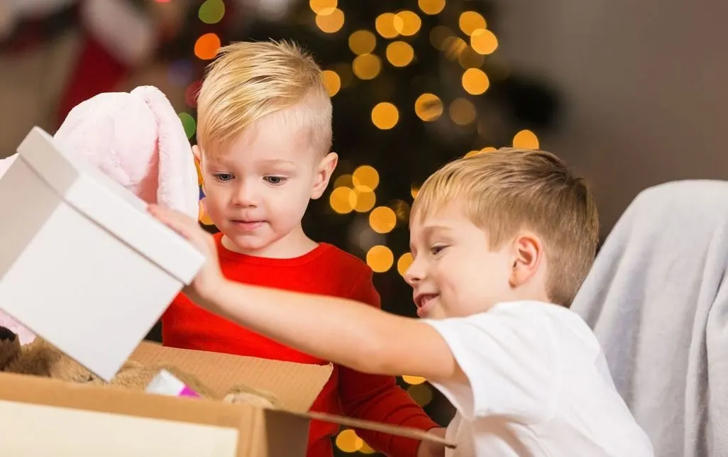 Zwei Kinder packen gemeinsam eine Geschenkbox vor einem Weihnachtsbaum aus