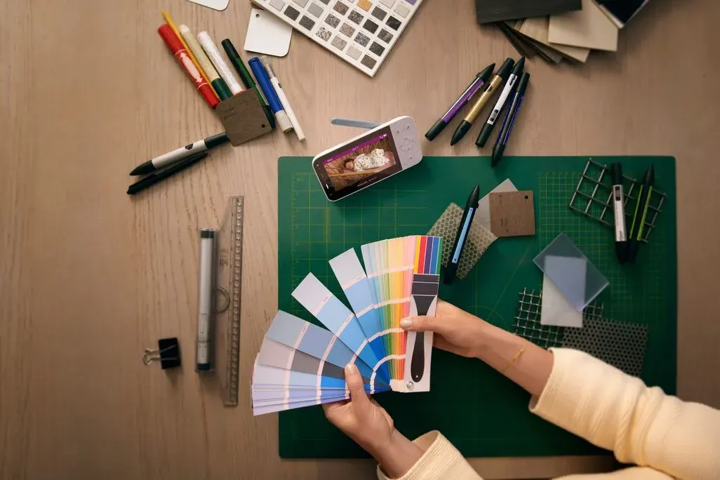 Blick von oben auf einen Schreibtisch mit Zeichen‑ und Bastelmaterialien, in dessen Mitte eine Person Farbmusterkarten auffächert, während daneben ein Babyphone mit Farbdisplay steht, auf dem ein schlafendes Baby zu sehen ist.