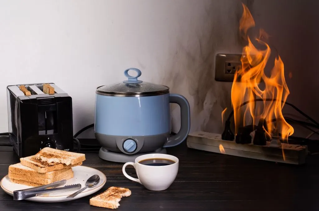 Toaster, Wasserkocher, ein Teller mit Toastbroat und eine Tasse Kaffee stehen neben einer brennenden Steckdosenleiste.