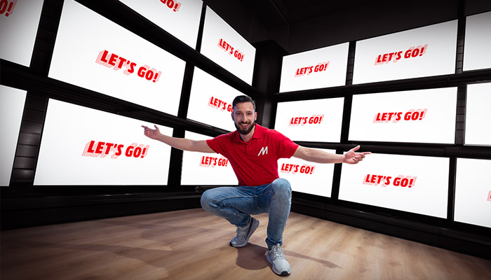 Ein Raum mit Holzboden und mehreren Bildschirmen mit dem roten Text "LET'S GO!".