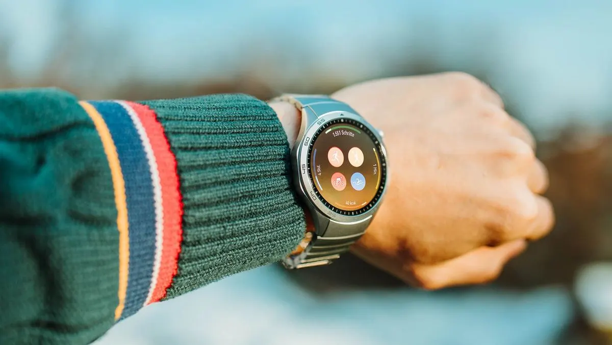Person mit grüner Strickjacke trägt eine runde Smartwatch mit Fitnessdaten auf dem Display.