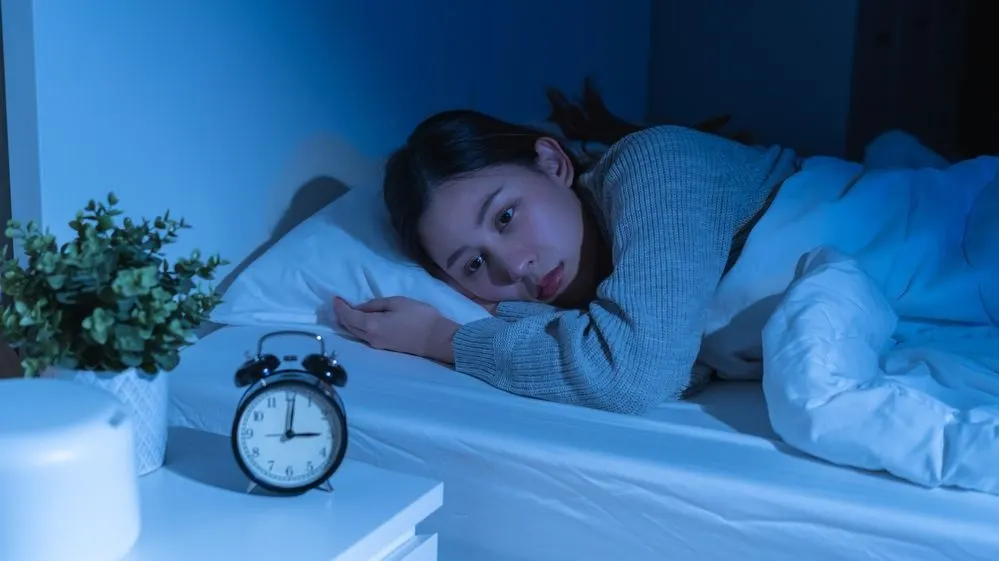 Person in Grau liegt wach im Bett. Schwarzer Wecker und grüne Pflanze auf weißem Nachttisch im blauen Raum.