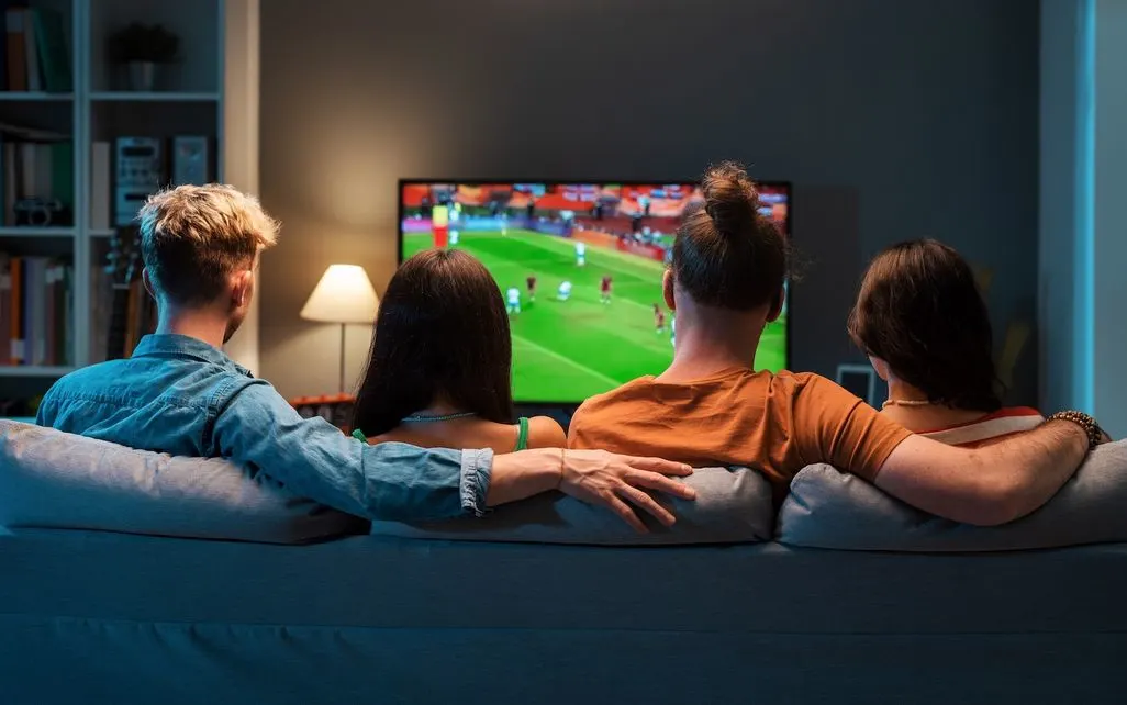 Vier Personen sitzen auf einem Sofa und schauen gemeinsam Fußball im Fernsehen.