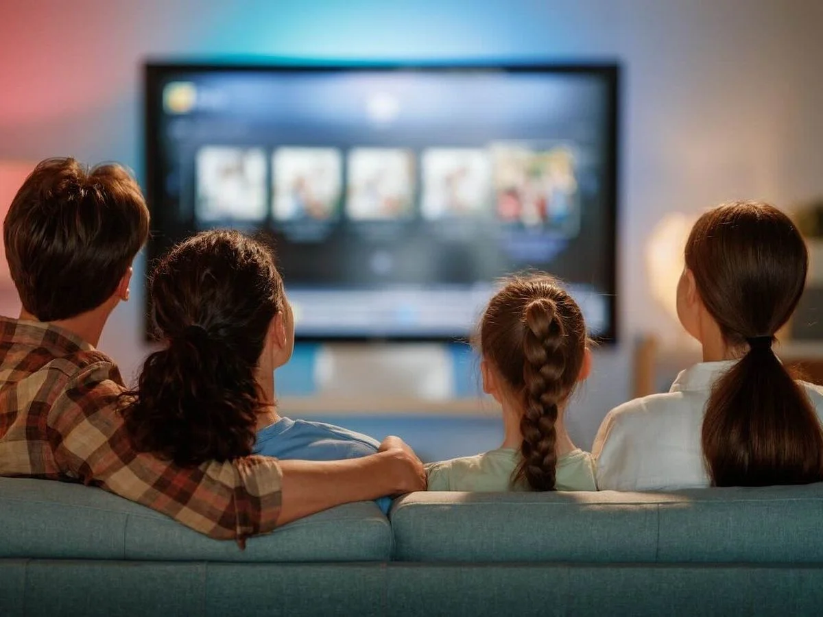 Vier Personen sitzen auf einem blaugrauen Sofa und blicken auf einen verschwommenen Smart-TV mit Thumbnails. Eine Person umarmt eine andere. Die Wand ist farbig beleuchtet.