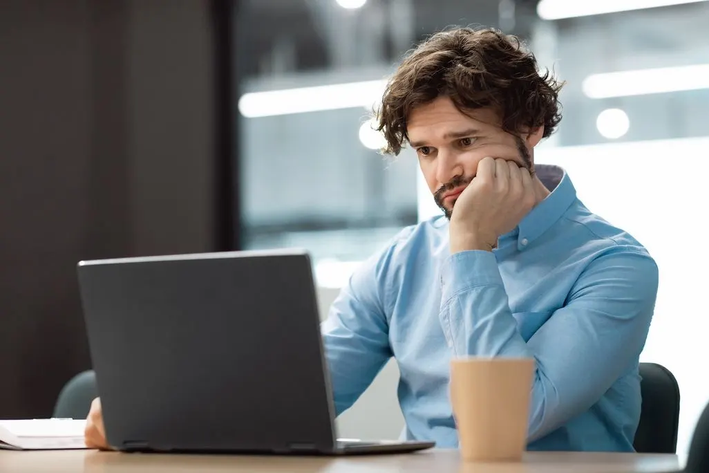 Mann in blauem Hemd sitzt nachdenklich vor Laptop im modernen Buero