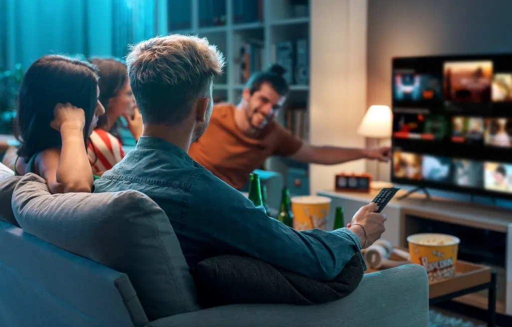 Gruppe von Freunden sitzt auf Sofa mit Popcorn und Getraenken und waehlt einen Film am Fernseher