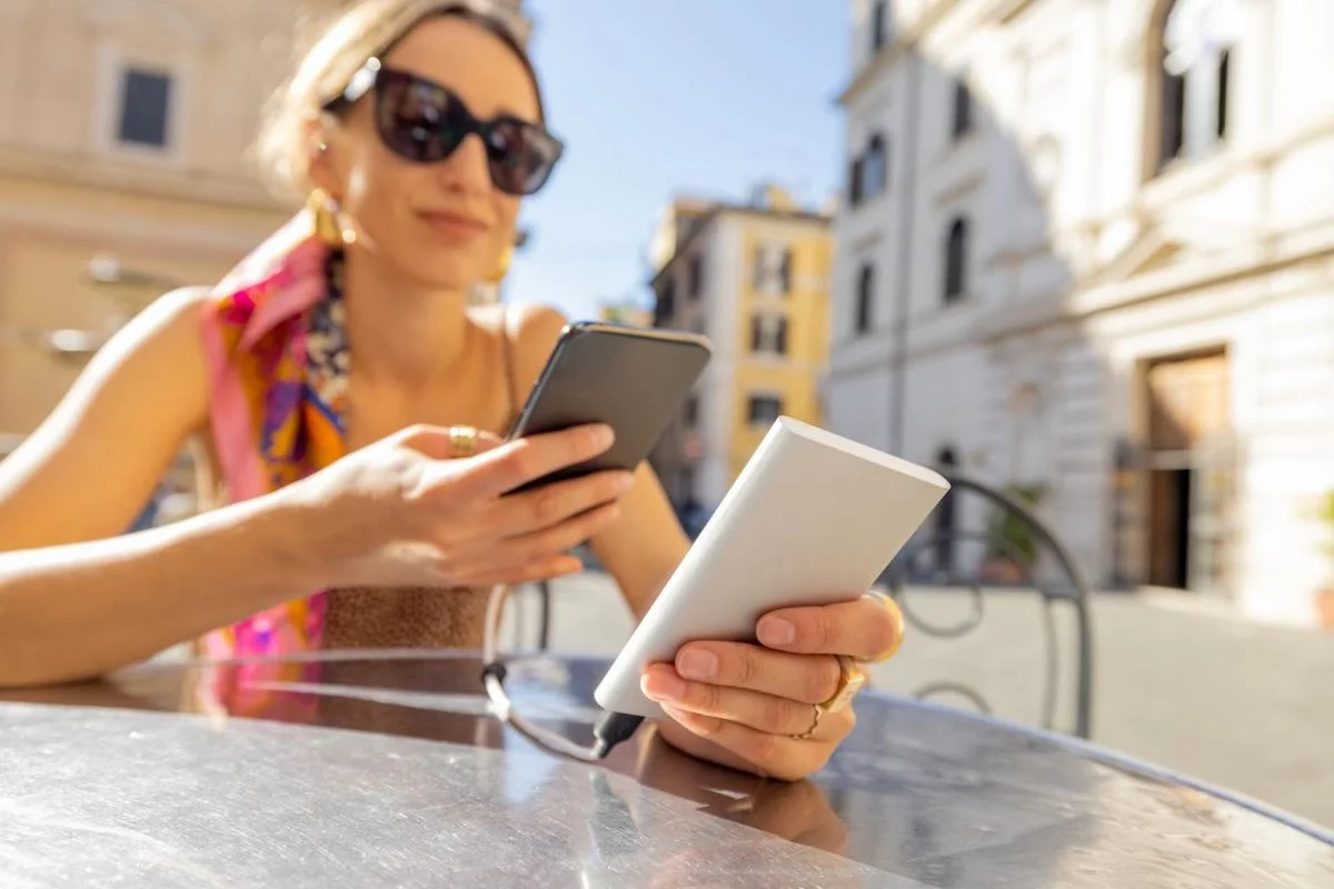 Frau mit Sonnenbrille sitzt in einer südeuropäischen Altstadt und hält zwei Tablets oder mobile Geräte in der Hand