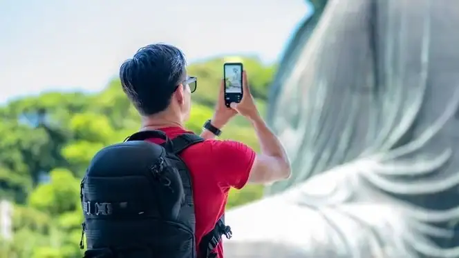 Person mit rotem Hemd, schwarzem Rucksack, Sonnenbrille und Uhr, fotografiert mit schwarzem Smartphone.